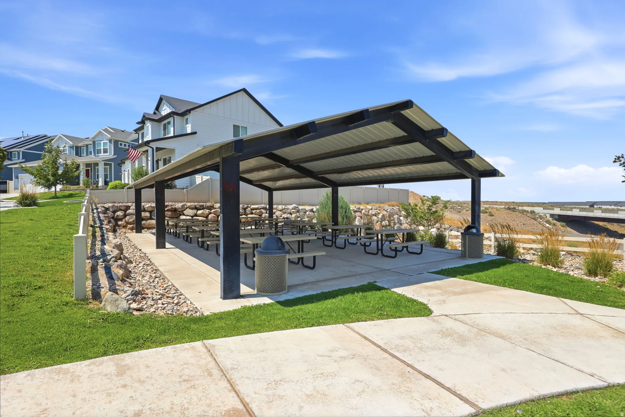 Community picnic pavilion featuring a metal roof, concrete slab, multiple picnic tables, and a stone retaining wall