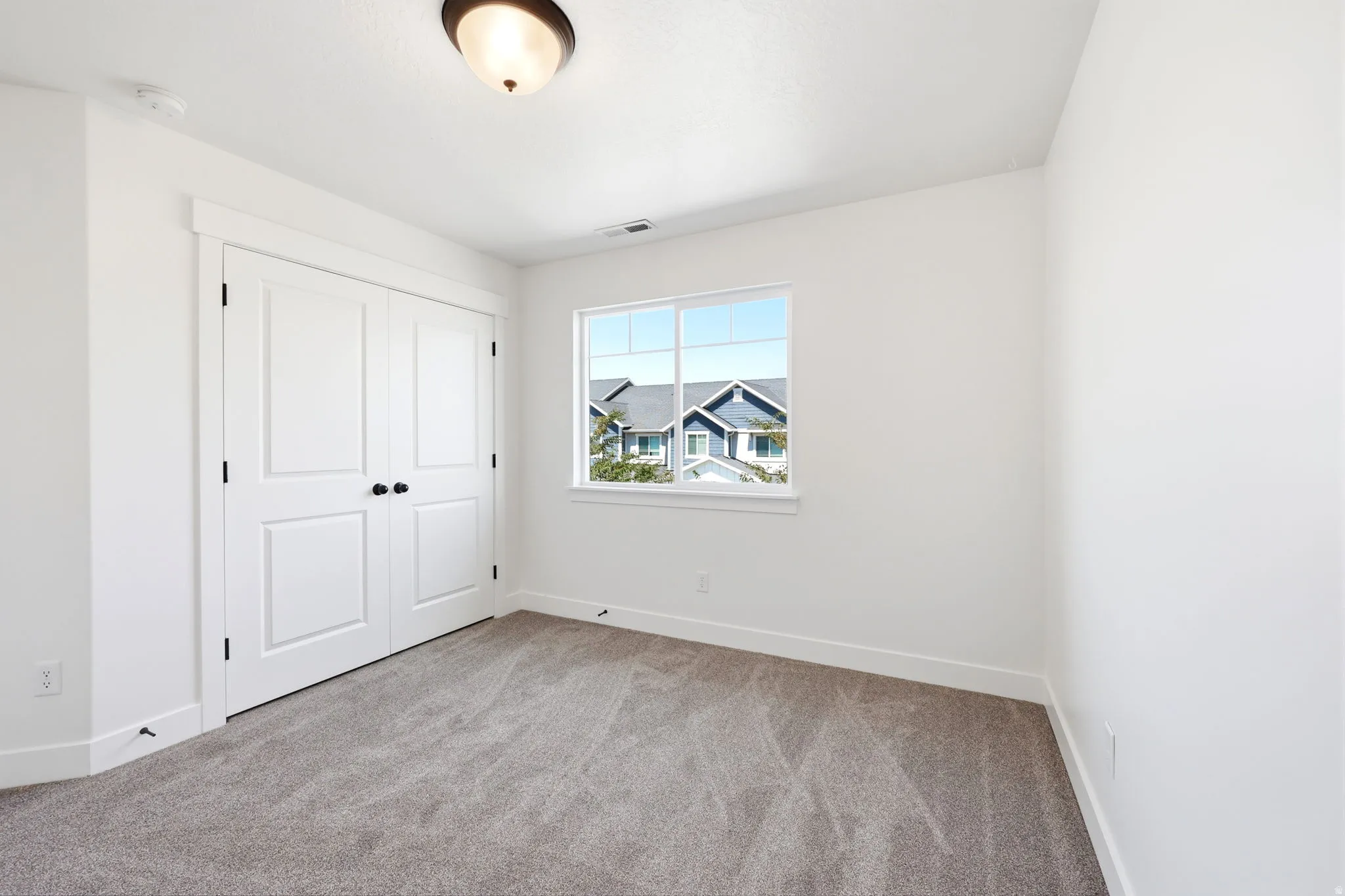 Bright room featuring a window with exterior views, light gray carpeting, crisp white walls, white baseboards, and a double-door closet with black hardware