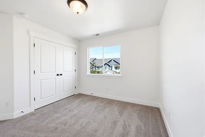 Bright room featuring a window with exterior views, light gray carpeting, crisp white walls, white baseboards, and a double-door closet with black hardware