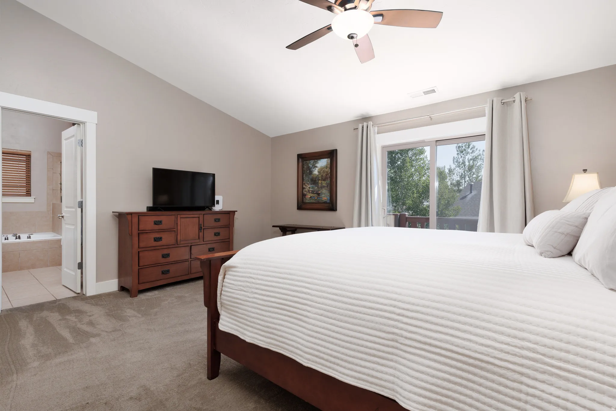 Vaulted ceiling with a ceiling fan, sliding glass door, plush carpet flooring, and an open doorway to a bathroom with a built-in tub
