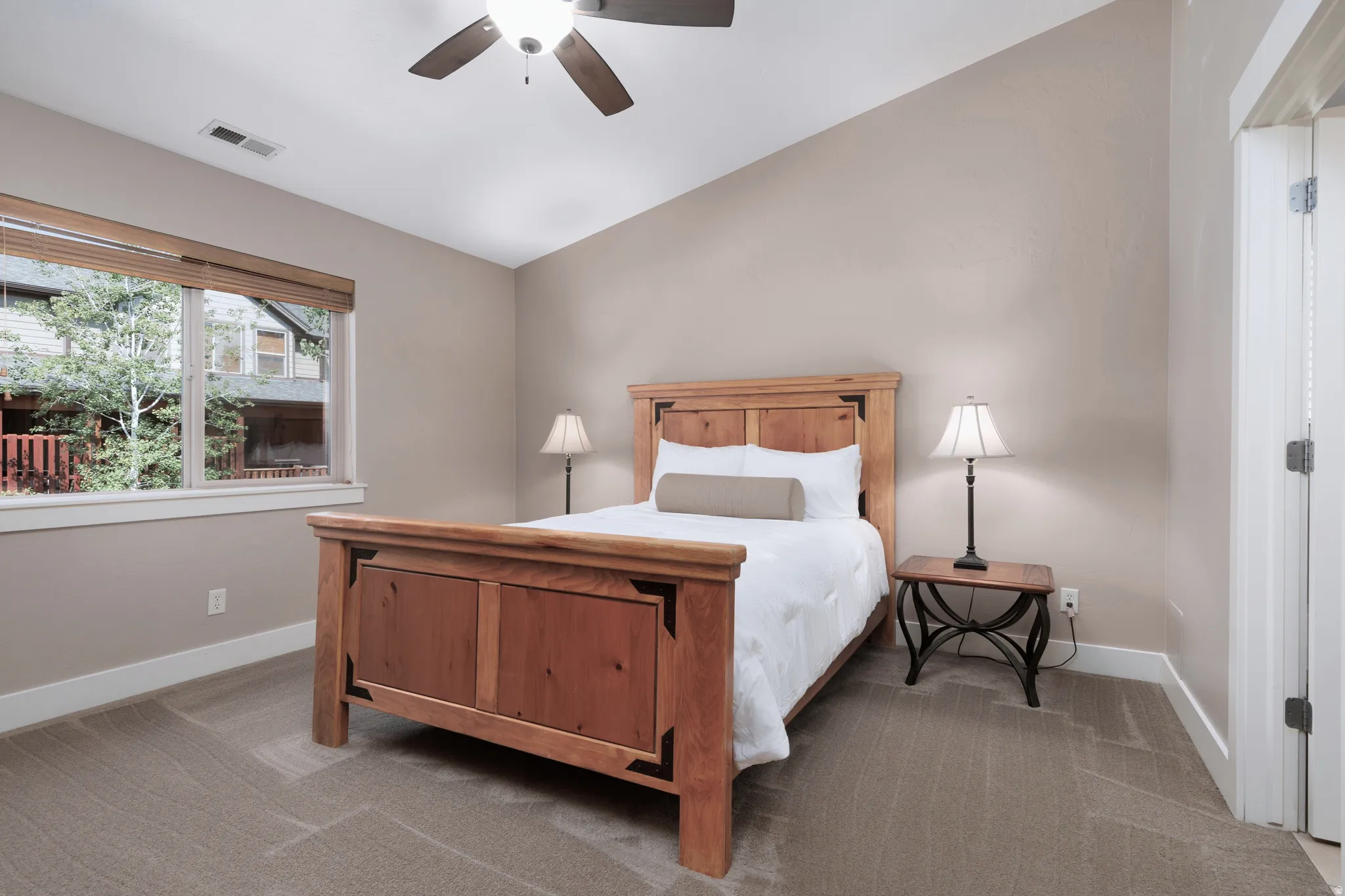 Vaulted ceiling, ceiling fan, large window with blinds, neutral wall tones, and plush carpeting