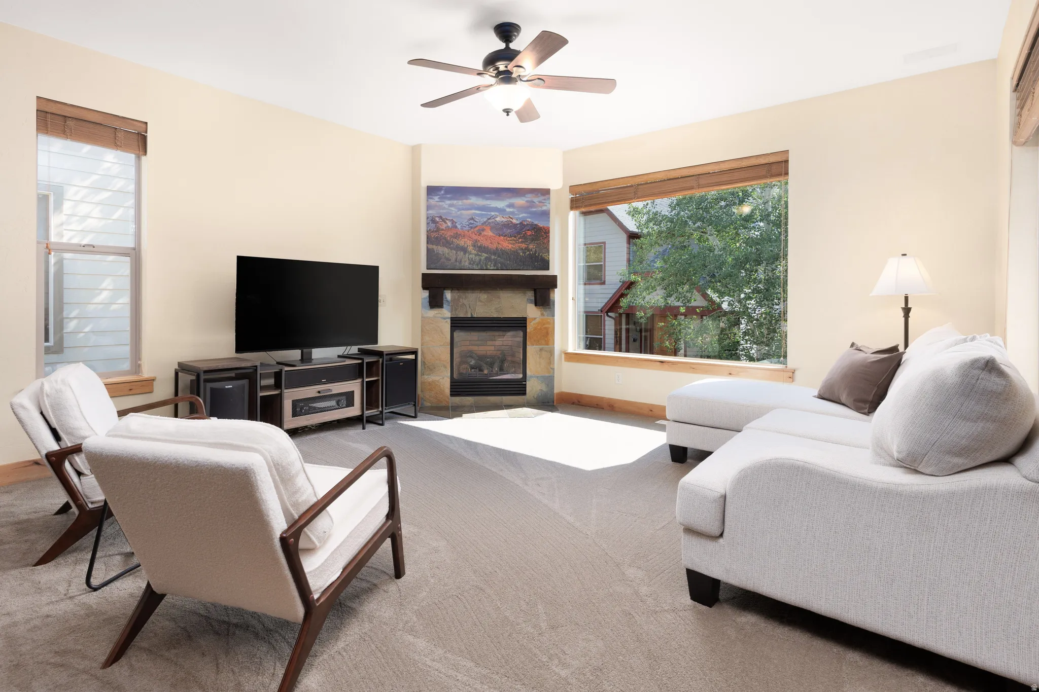 Corner stone-clad fireplace with wood mantle, ceiling fan with light fixture, large picture window with wood trim, additional tall window with wood trim, and light-colored wall paint