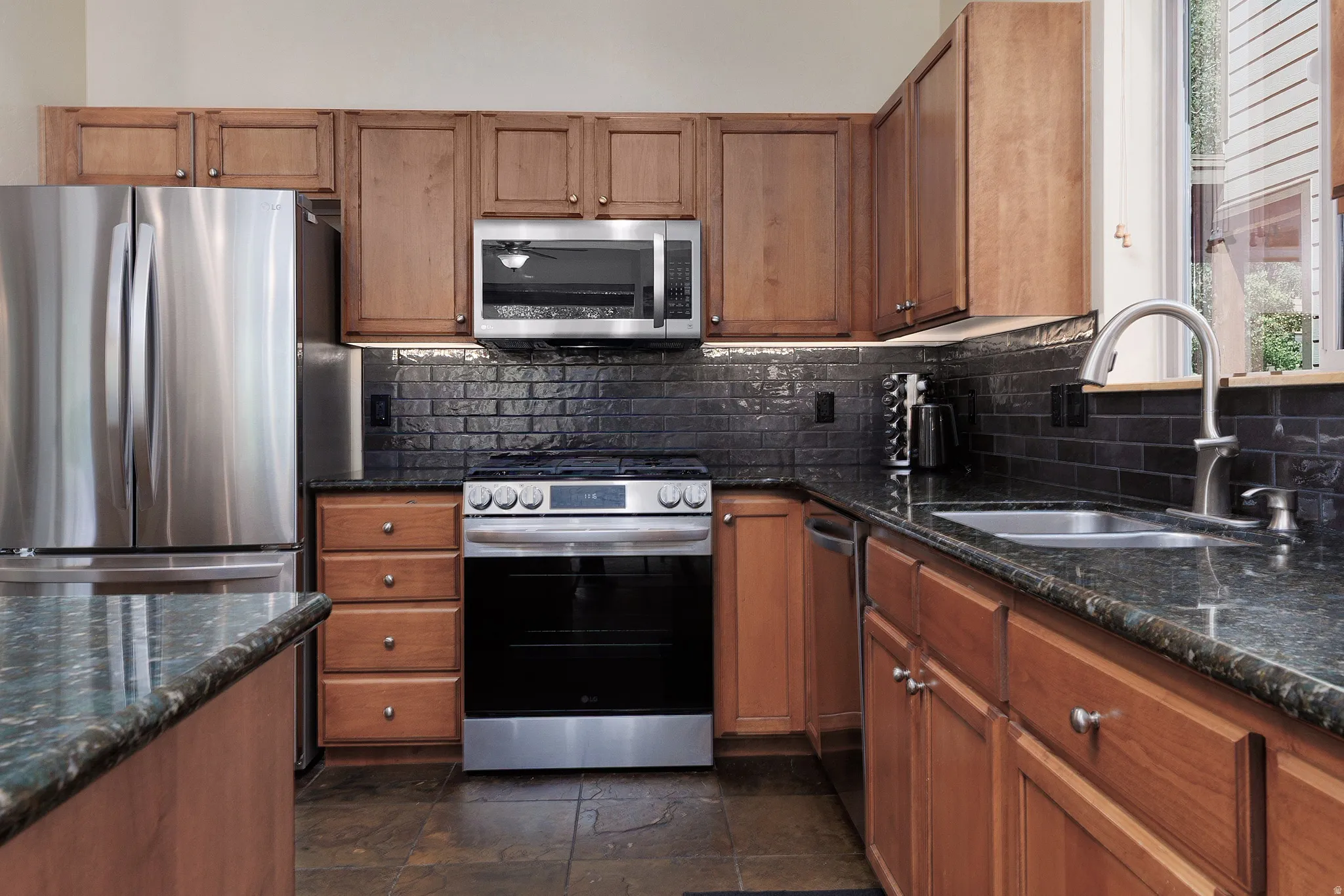 Wood cabinetry with stainless steel hardware, black subway tile backsplash, dark-toned countertops with an integrated sink, and stainless steel appliances