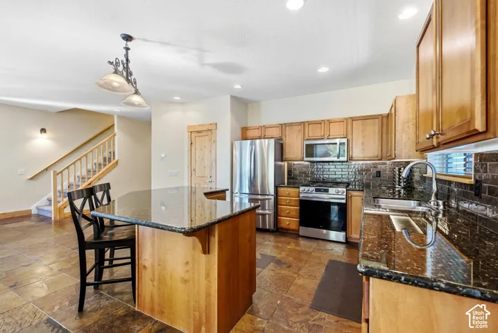 Kitchen featuring an island with a dark countertop, stainless steel appliances, wood cabinetry, and a dark tile backsplash
