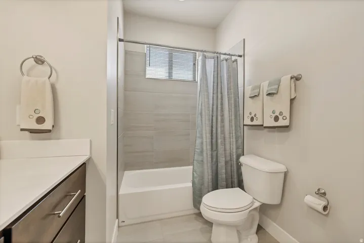 White bathtub with light gray tile surround, window with blinds, and light-colored flooring