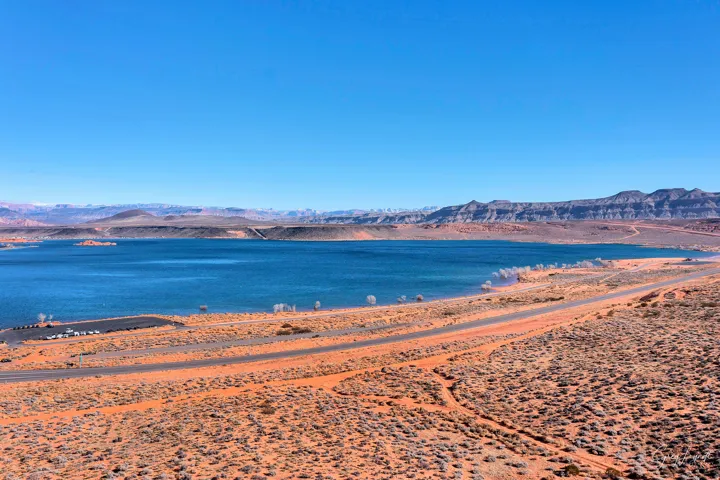 Expansive blue lake with a mountainous backdrop, bordered by a desert landscape, and a paved road
