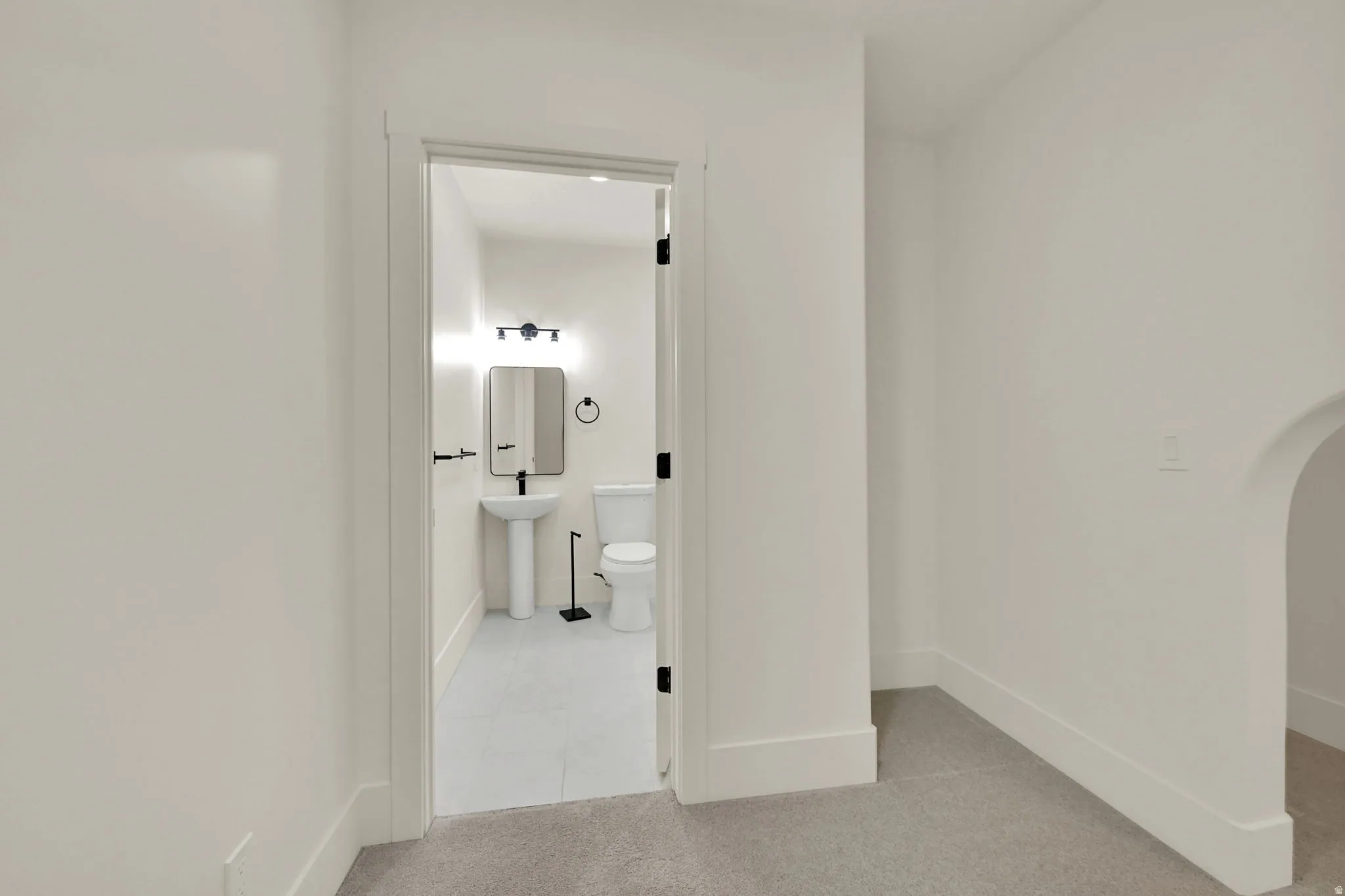 White pedestal sink, modern wall-mounted light fixture, rectangular mirror, toilet, and light-toned flooring
