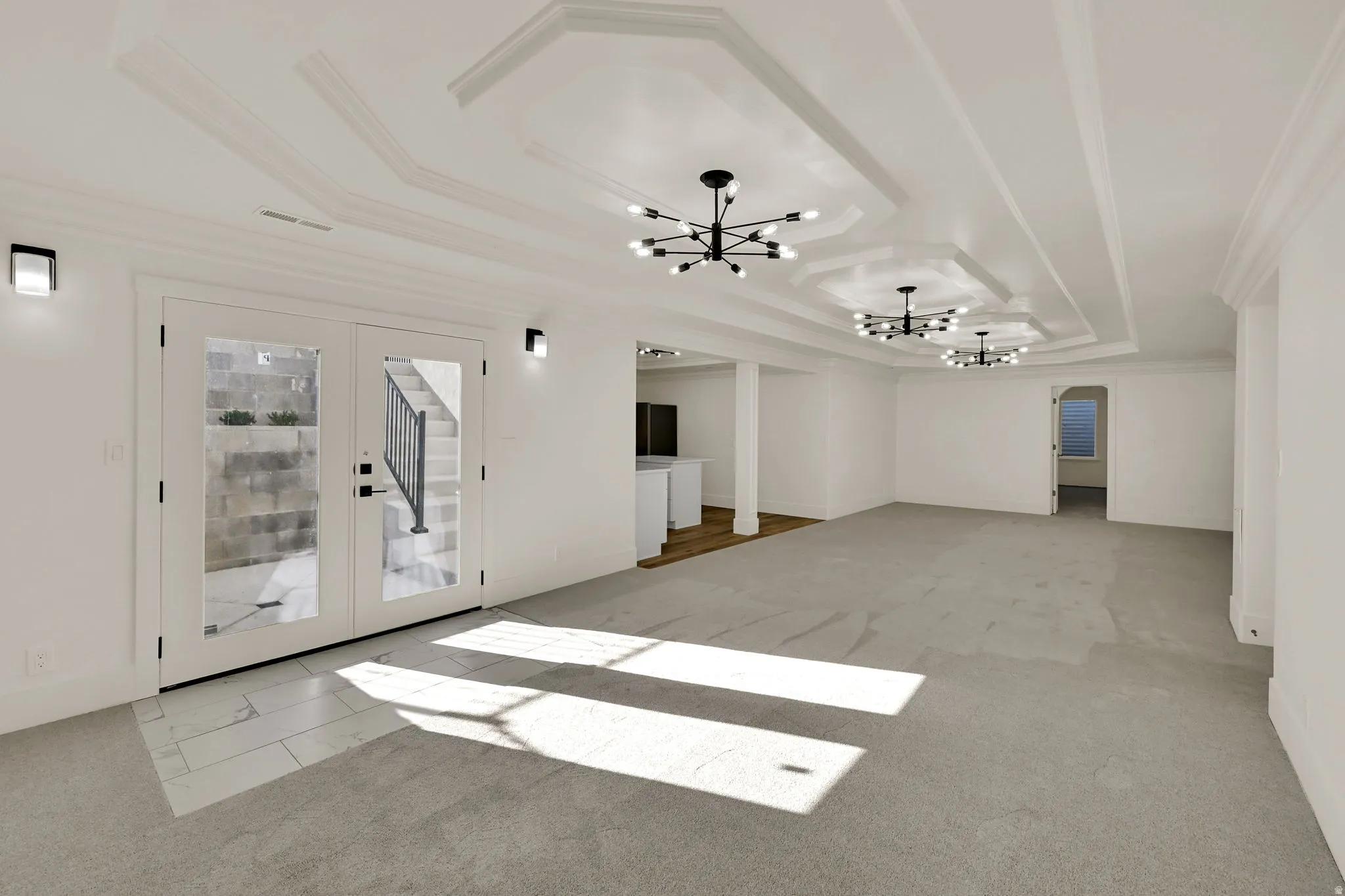 Recessed tray ceilings with contemporary light fixtures, double French doors with transom windows, wall-mounted sconces, and mixed flooring with carpet and tile