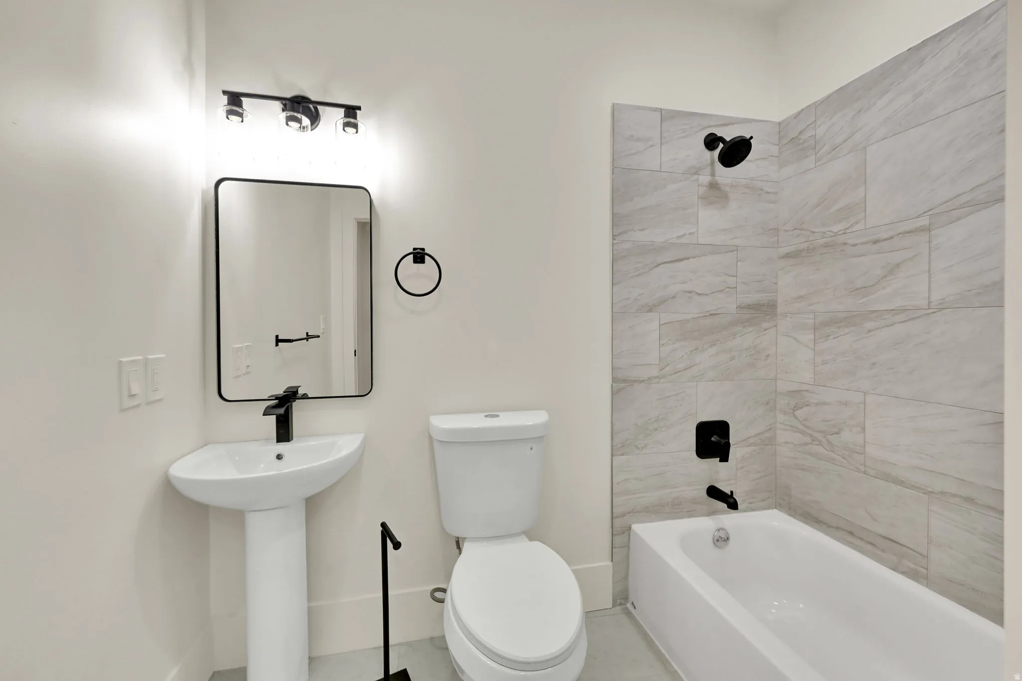 Pedestal sink with a matte black faucet, rectangular mirror with a black frame, track lighting with three fixtures, white bathtub, and light gray tile shower surround