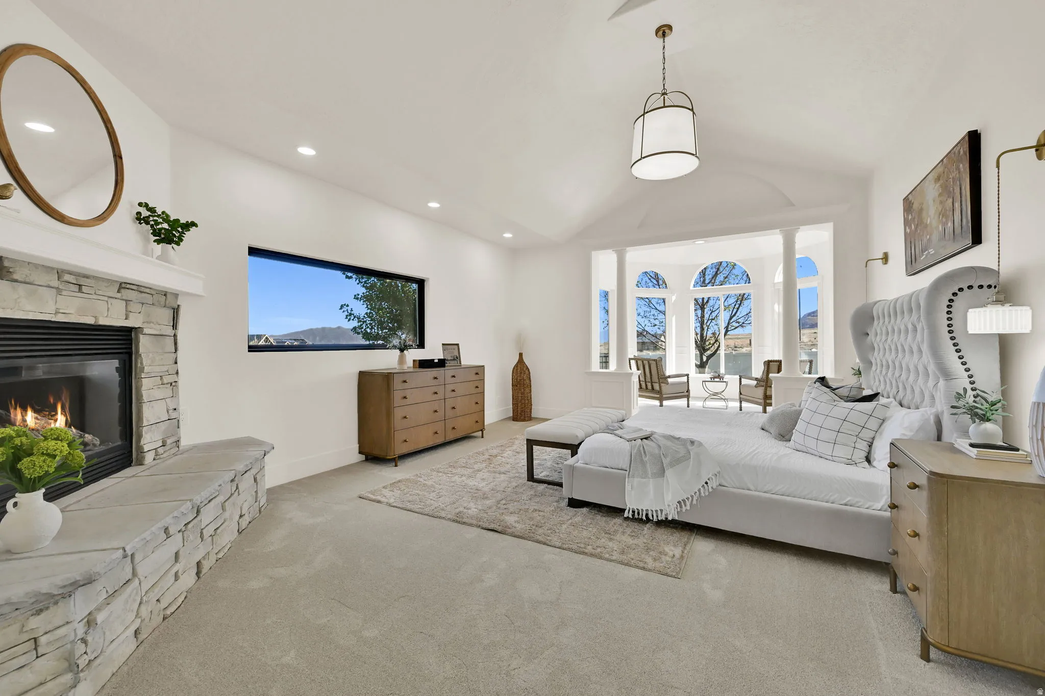 Primary Suite with vaulted ceiling with pendant lighting, stone-clad fireplace, picture window, and bay window with arched transoms