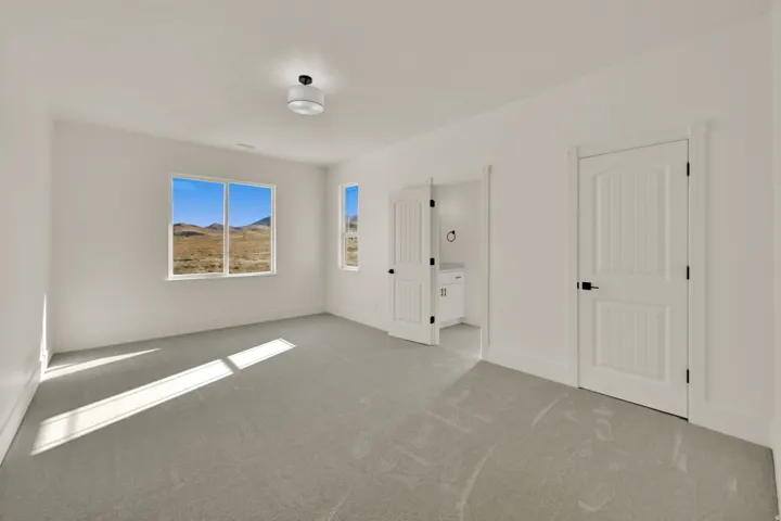 Spacious room featuring light gray carpeting, crisp white walls, multiple windows with a natural landscape backdrop, and white paneled doors with black hardware