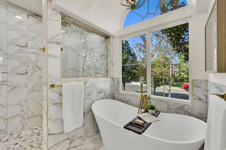 Primary Suite with freestanding soaking tub, walk-in shower with glass enclosure, marble-style wall tiling, pebble shower floor, and arched transom window
