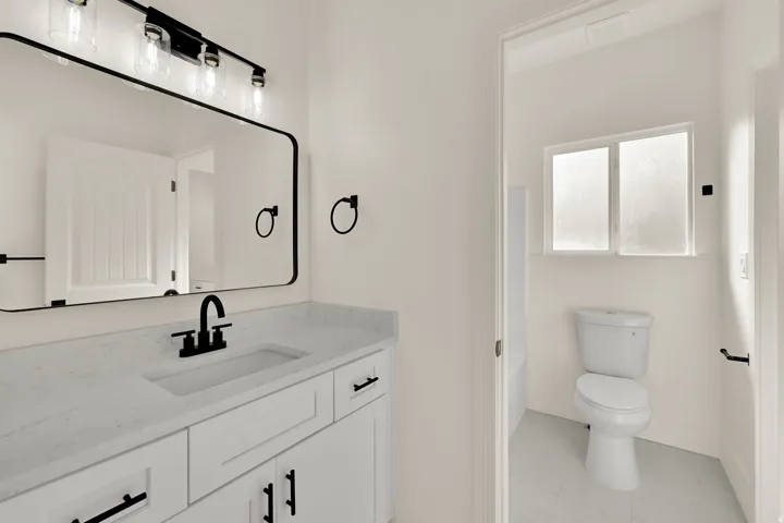White vanity with integrated rectangular sink and matte black faucet, framed mirror with black trim, black vanity light fixture, and white wall panels