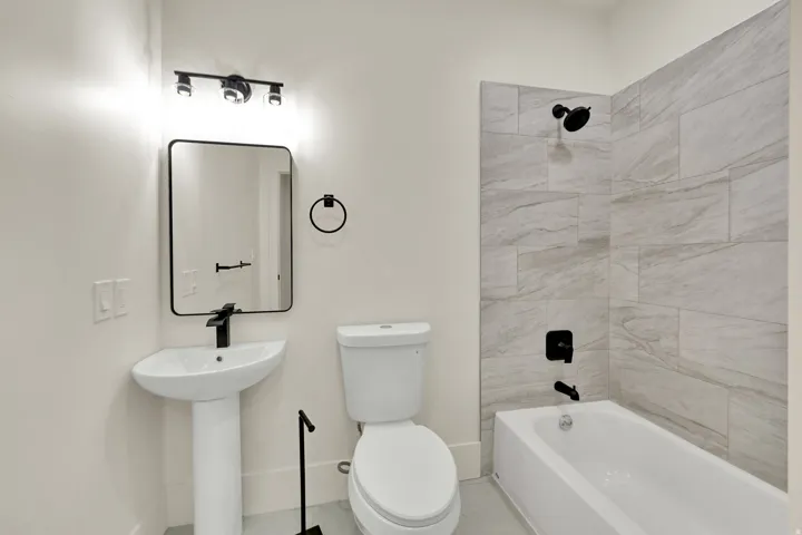 Pedestal sink with a matte black faucet, rectangular mirror with a black frame, track lighting with three fixtures, white bathtub, and light gray tile shower surround