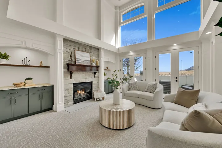 Two-story windows, stone-clad fireplace with a wood mantel, built-in cabinetry with stone countertops, recessed lighting, and double French doors leading to an exterior space