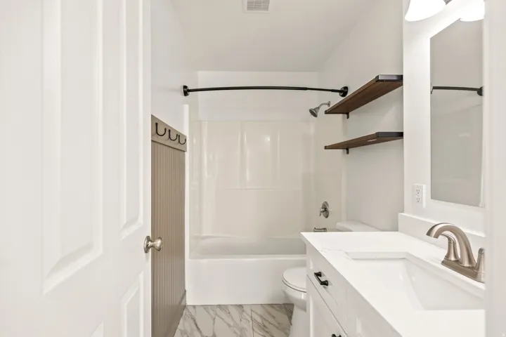 Bathroom featuring a white vanity with an integrated sink, brushed nickel faucet, wood-finish shelving, a white bathtub with shower, and marble-patterned floor tiles
