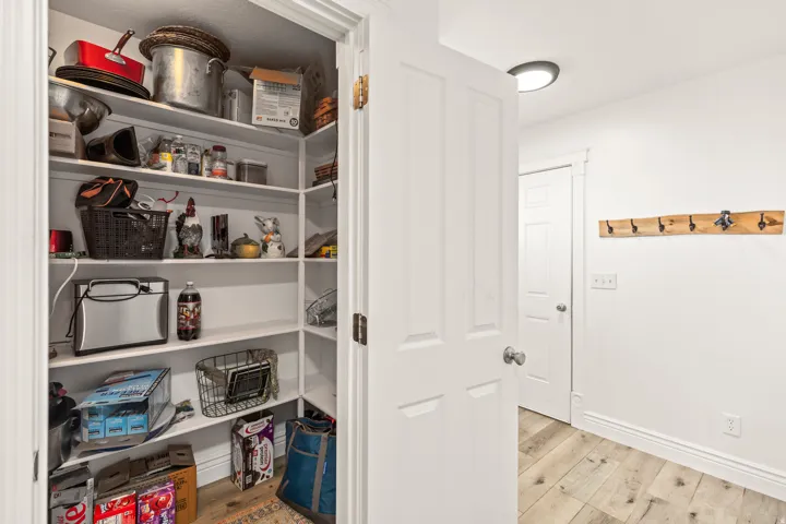Walk-in pantry with ample shelving, white panel door, light wood-finish flooring, and a wall-mounted coat rack