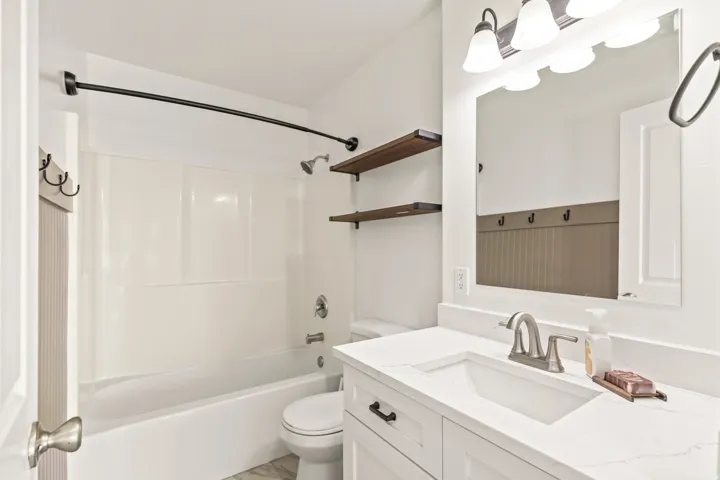 White vanity with a rectangular sink and brushed nickel faucet, framed mirror with a three-light fixture, tub-shower combination with a curved rod, and wood-finish floating shelves