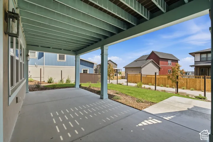 Covered outdoor patio with concrete flooring, supported by structural pillars, featuring exterior lighting fixtures, a manicured lawn, and perimeter fencing