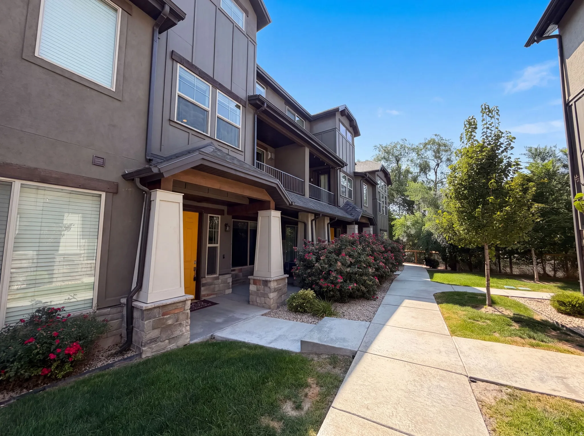 Modern townhome exterior featuring a covered entry, stone column bases, a bright yellow entry door, and a concrete walkway bordered by landscaping