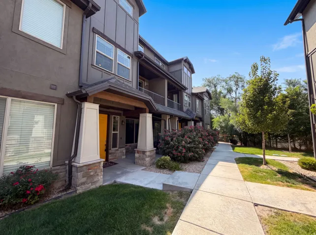Modern townhome exterior featuring a covered entry, stone column bases, a bright yellow entry door, and a concrete walkway bordered by landscaping