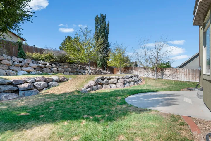 Curved concrete patio with a tiered rock retaining wall, featuring a wood fence, established trees, and natural grass
