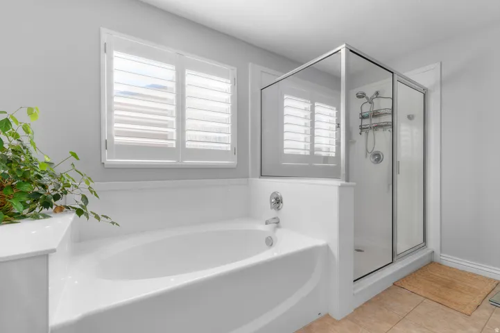 Soaking tub with chrome fixtures, glass-enclosed shower, white plantation shutters, and light tile flooring