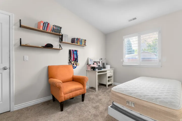 Vaulted ceiling, wall-mounted wood-finish shelving, desk with integrated storage, window with plantation shutters, and carpet flooring
