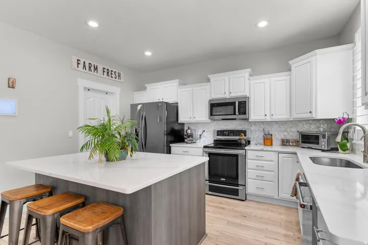 Central island with a light-toned countertop, white cabinetry with crown molding, stainless steel appliances, patterned tile backsplash, and light wood-finish flooring