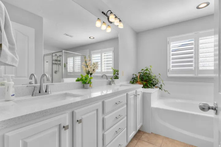 Dual sink vanity with white cabinetry, built-in bathtub, and plantation shutters