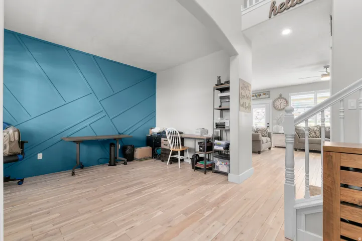 Wood-finish flooring throughout, accent wall with geometric paneling, white painted staircase with wooden newel post, recessed lighting, and a ceiling fan in the background