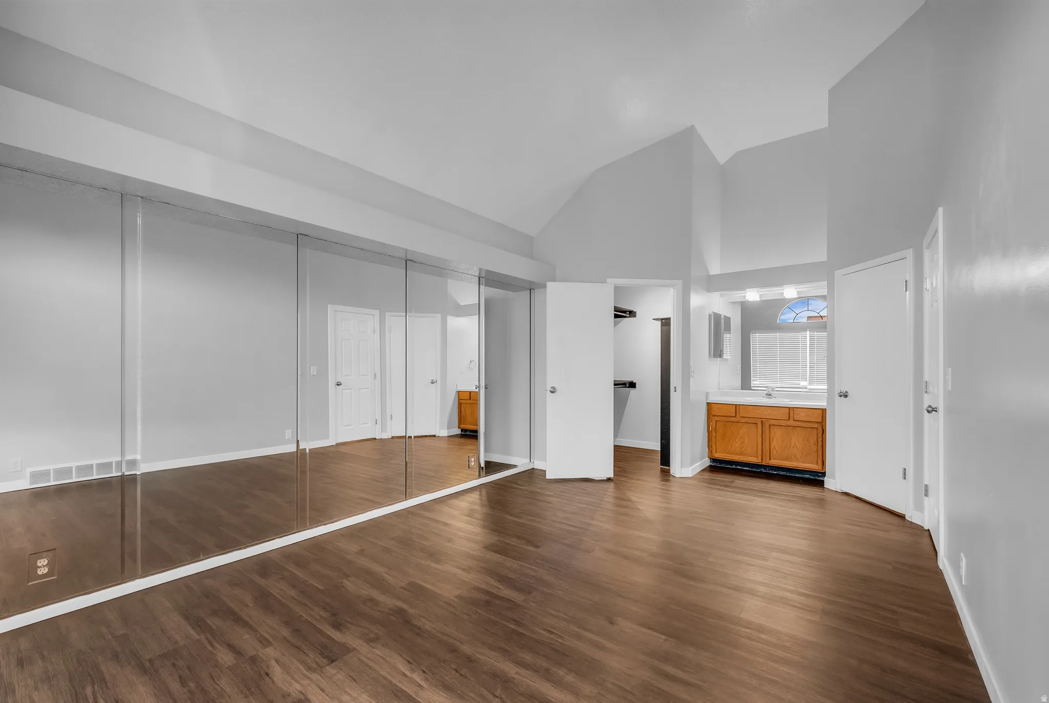 Vaulted ceilings, wood-finish flooring, mirrored closet doors, built-in vanity with wood cabinetry, and an arched window with blinds