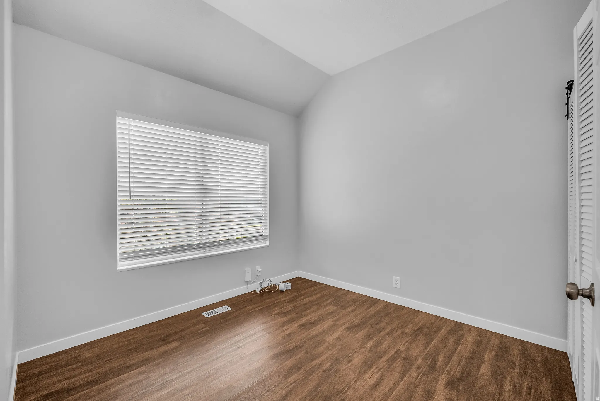 Sloped ceiling room featuring wood-finish flooring, a window with horizontal blinds, and a louvered door