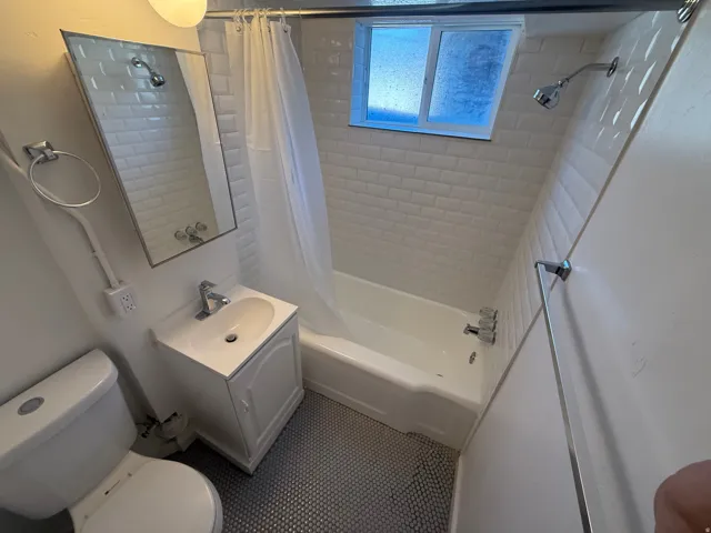 White subway tile shower surround, window with privacy glass, white bathtub, white vanity with integrated sink, and dark penny tile flooring