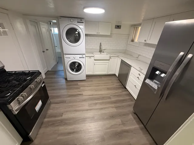 Full kitchen with stainless steel appliances, white subway tile backsplash, white shaker cabinetry, wood-finish flooring, and stacked laundry machines