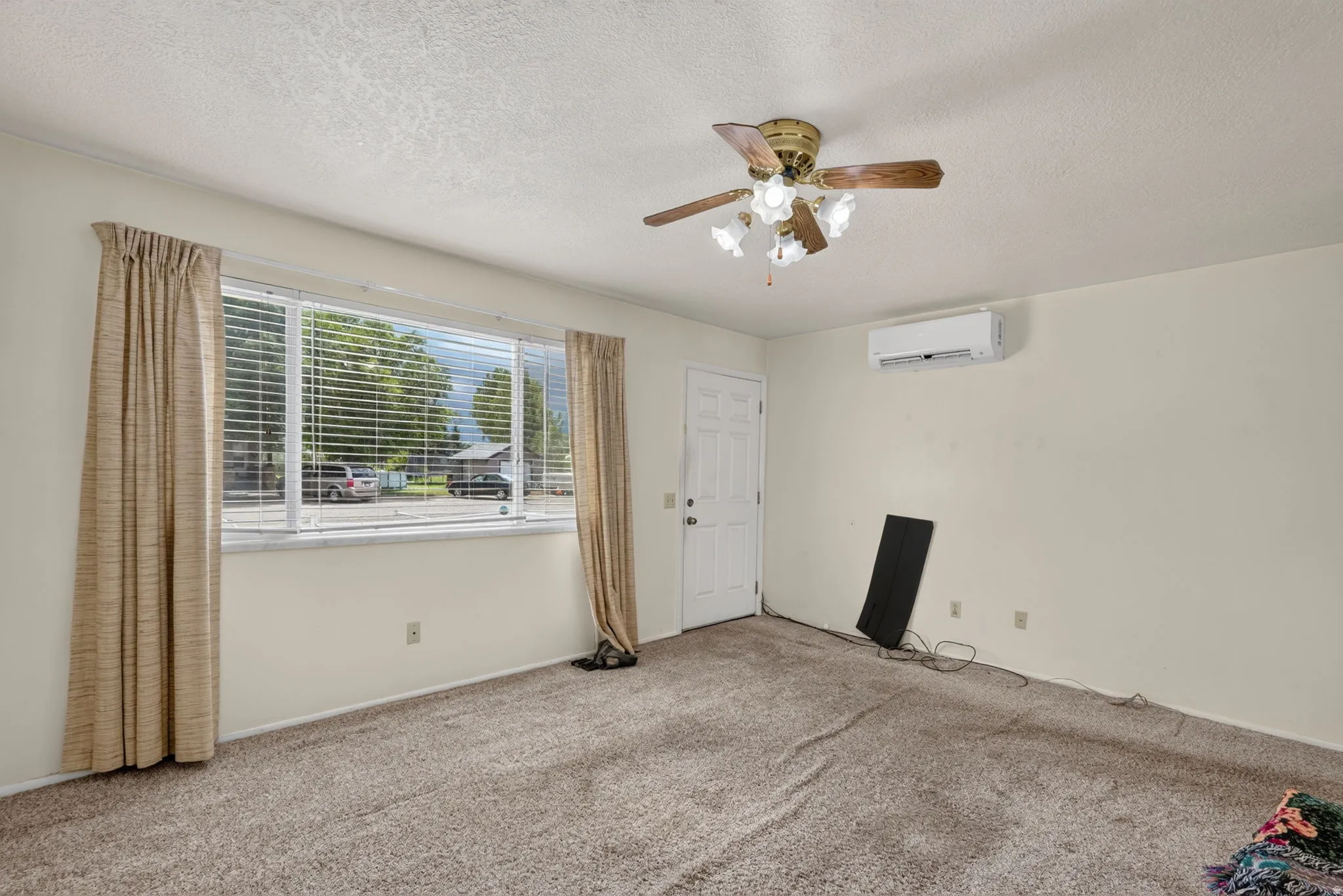 Wall-mounted air conditioning unit, ceiling fan with light fixture, broad window, textured carpeting, and white paneled door