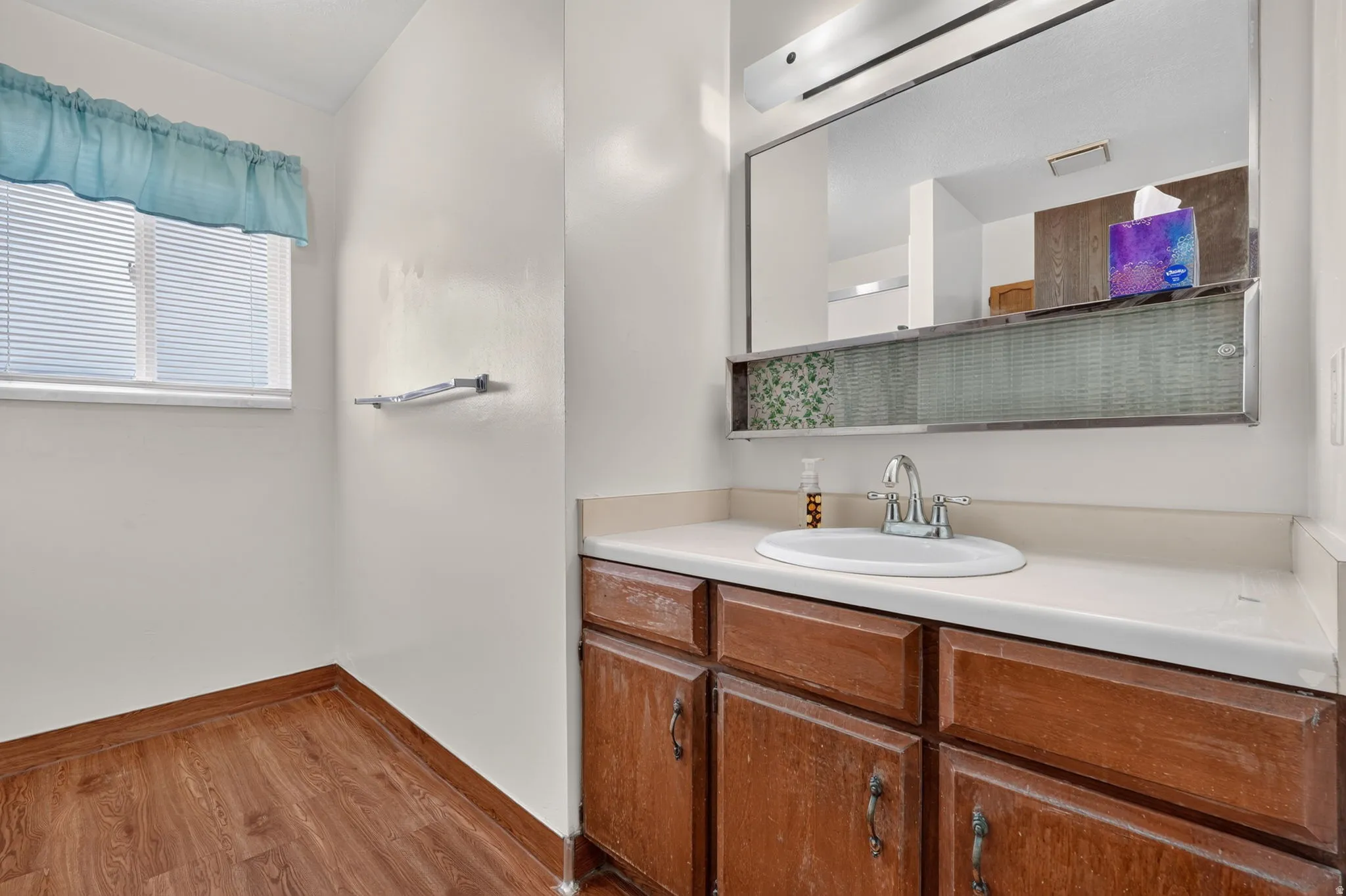 Wood-finish flooring, single vanity with integrated sink, window with horizontal blinds, built-in mirror with integrated lighting, and towel rack