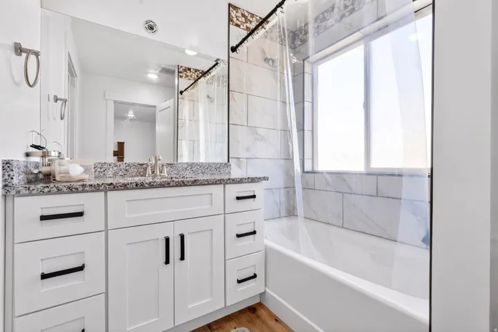 White vanity with black hardware, stone-finish countertop, wood-finish flooring, and white subway tile bath surround