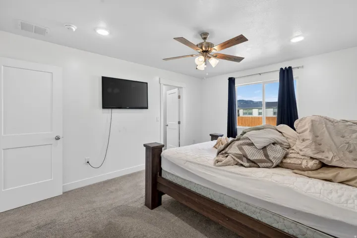 Master bedroom featuring a wood-finish bed frame, carpet flooring, ceiling fan with integrated lighting, wall-mounted flat-screen television, and expansive windows with mountain vistas and blue drapery