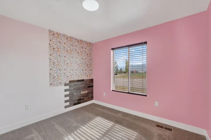 Bright bedroom with fresh pink accent walls, large window with horizontal blinds overlooking pastoral landscape, wood-finish flooring, and a decorative tile accent wall featuring warm earth-tone mosaic patterns
