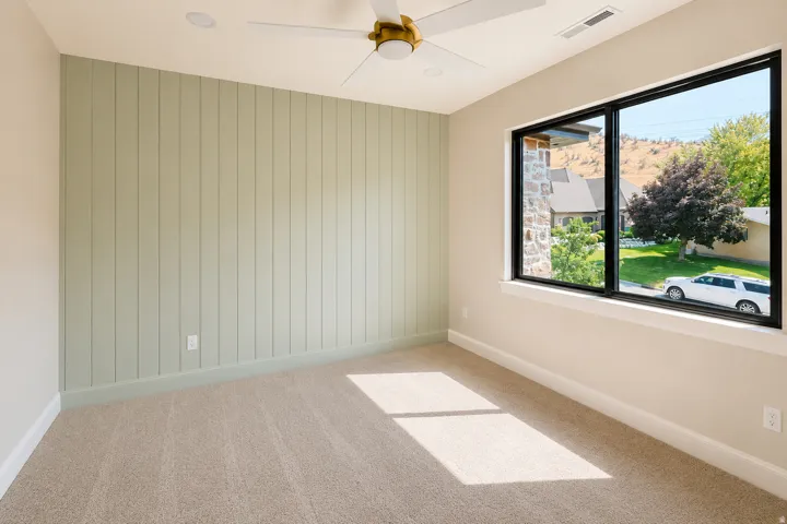 Bedroom 2 with an accent wall with vertical paneling, new carpet, windows, and a ceiling fan.