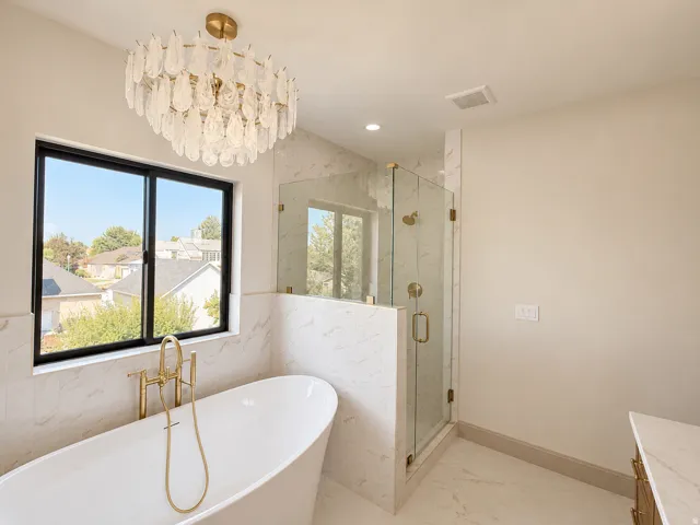 Freestanding soaking tub with a gold-finish filler, walk-in shower with clear glass enclosure and gold-finish fixtures, and a beautiful chandelier