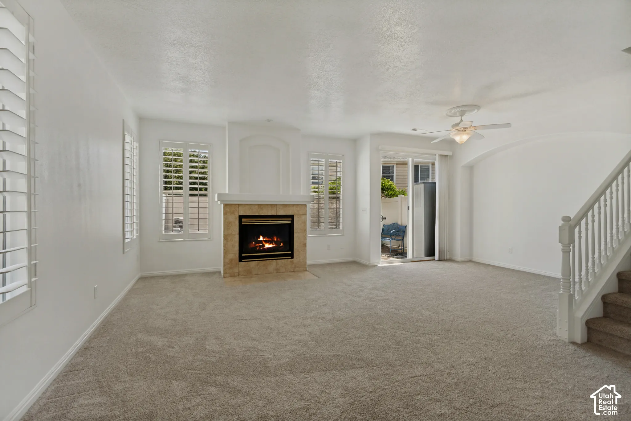 Living area featuring a fireplace with a stone tile surround, multiple windows with plantation shutters, ceiling fan, and carpeted flooring