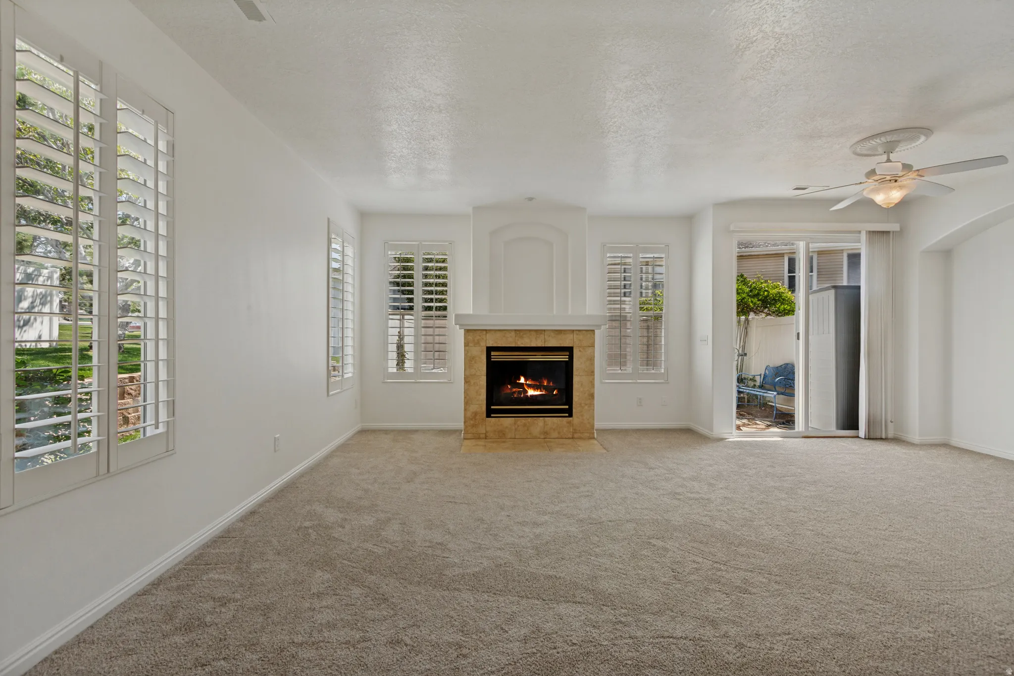 Carpeted living area featuring a fireplace with a tile surround, plantation shutters on multiple windows, a ceiling fan, and a sliding glass door