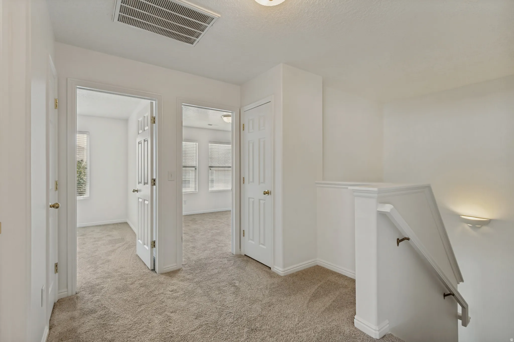 Carpeted hallway with white interior doors, a staircase with a white railing, and a wall-mounted light fixture