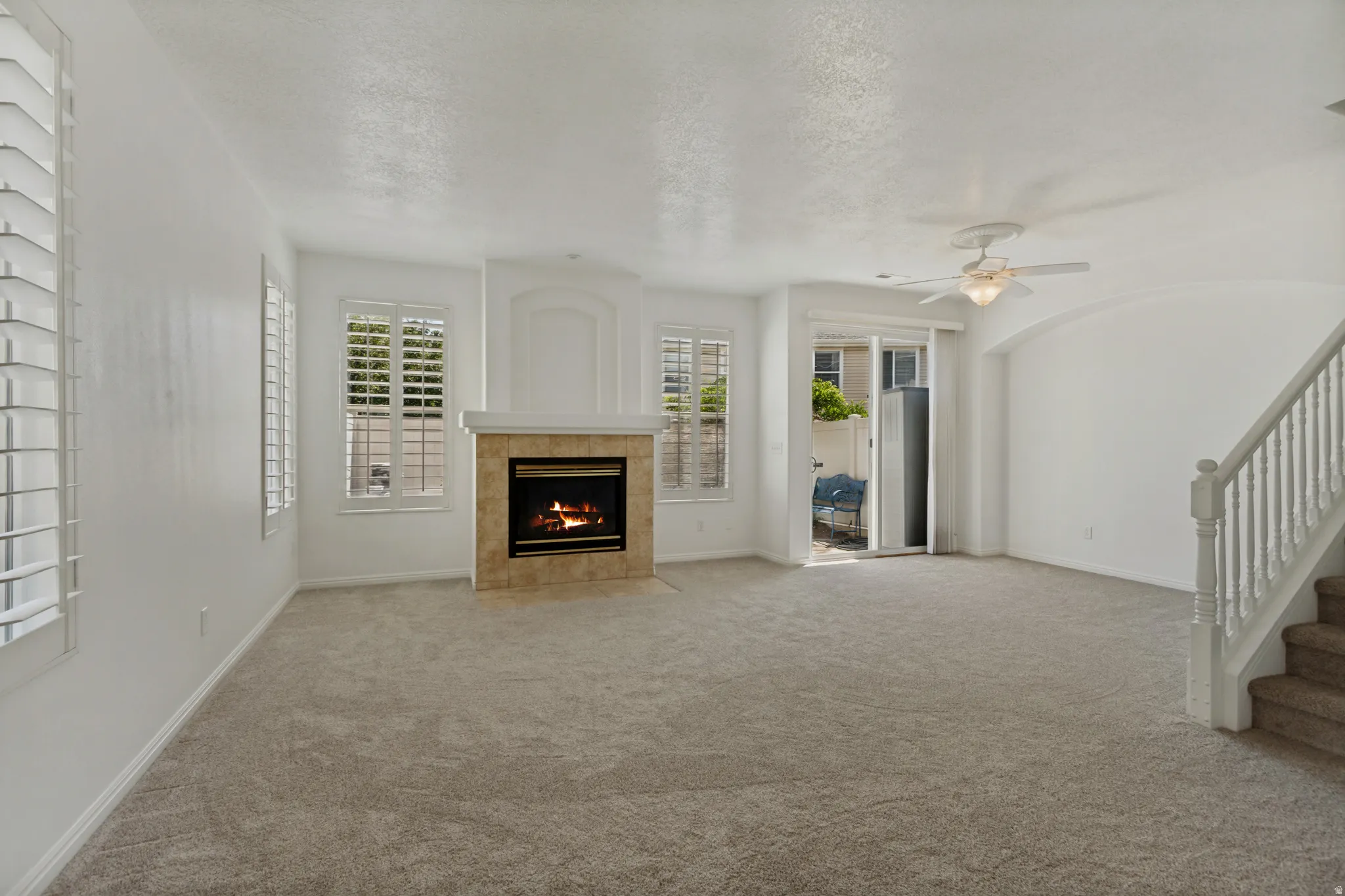 Living area featuring a fireplace with a stone tile surround, multiple windows with plantation shutters, ceiling fan, and carpeted flooring