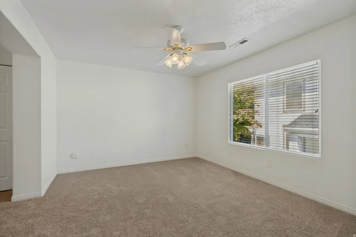Neutral-toned carpeting, white walls, ceiling fan with integrated lighting, and a large window with blinds
