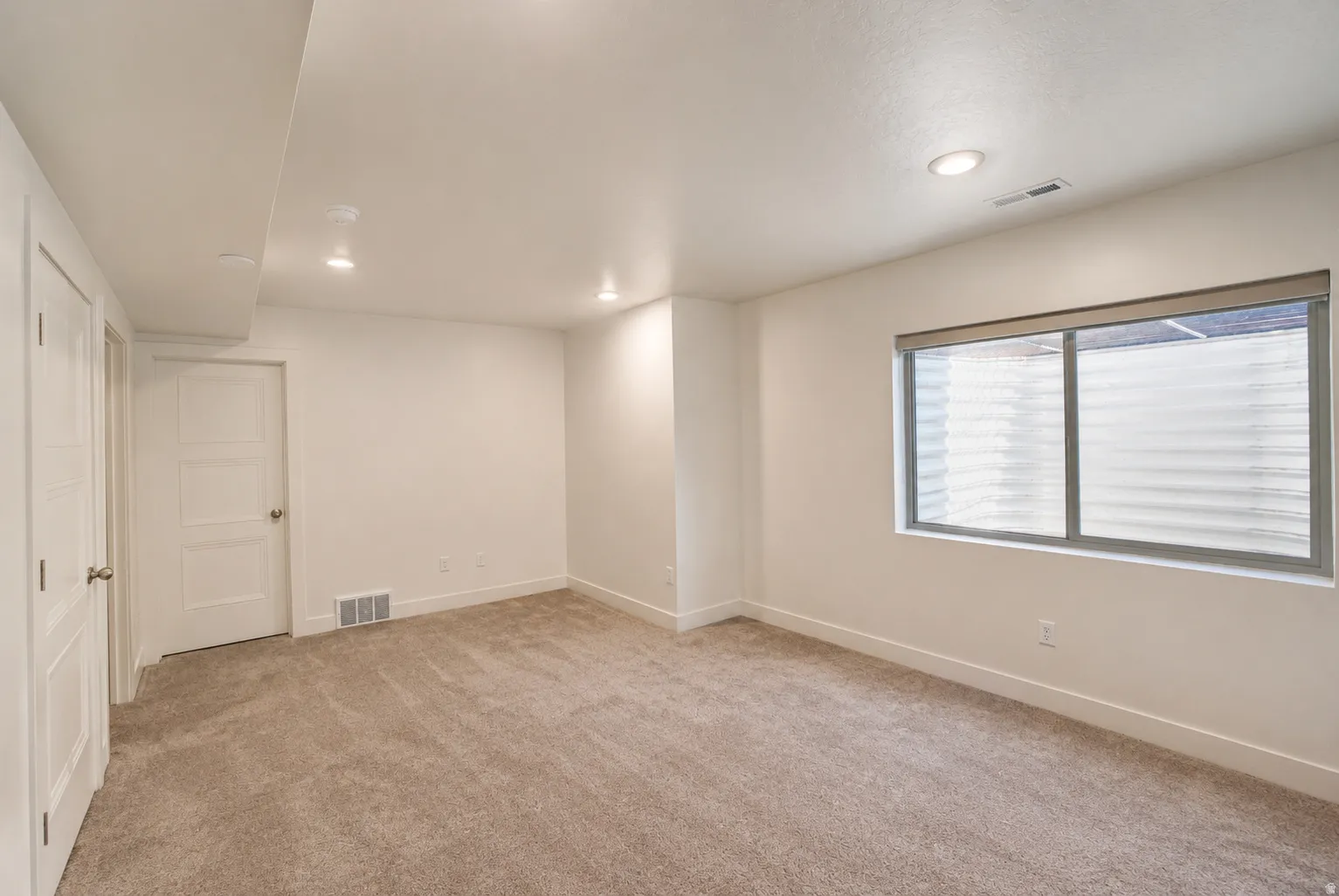 Recessed ceiling lighting, textured carpet flooring, white painted walls, and a large window with privacy blinds