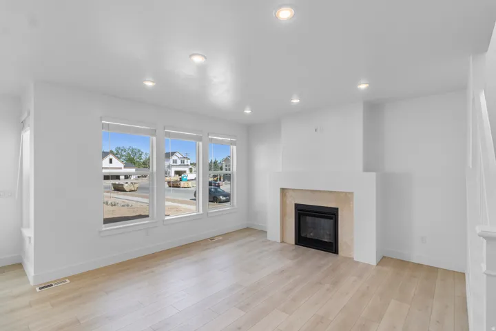Recessed lighting, light wood-finish flooring, three large windows, a fireplace with a light-toned tile surround, and a white mantel