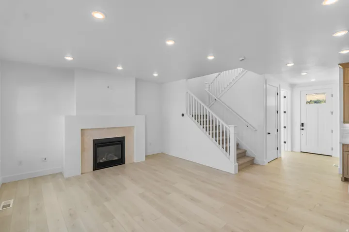 Recessed lighting throughout, a fireplace with a light-toned surround, light wood-finish flooring, a white staircase with wood railings, and a panel door with an upper glass insert